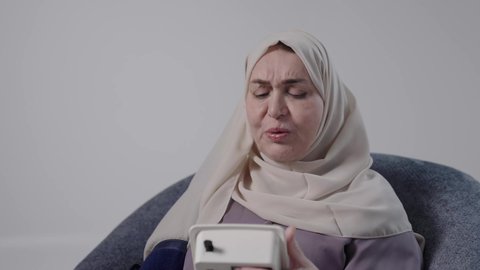 الاهتمام والرعاية بالصحة الجسدية, مفهوم الطب والرعاية الصحية, تصوير لامراه عربيه خليجيه سعوديه كبيره بالسن ترتدي العباءة والحجاب تجلس على كرسي ، تحمل جهاز الضغط وتقوم بقياسه ضغط دمها، الأجهزة الالكترونية المنزلية الحديثة والمتطورة