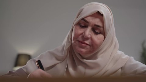 تدوين وتسجيل الملاحظات والمعلومات على الورق,  إدارة الأعمال وتنظيمها، تصوير لامراه عربيه خليجيه سعوديه كبيره بالسن ترتدي العباءه والحجاب تجلس على مكتب ، وتقوم بكتابة المهام والأهداف اليومية،  استخدام الاوراق البيضاء الفارغة في العمل