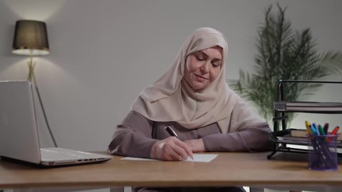 تدوين وتسجيل الملاحظات والمعلومات على الورق,  استخدام الاوراق البيضاء الفارغة في العمل، إدارة الأعمال وتنظيمها، تصوير لامراه عربيه خليجيه سعوديه كبيره بالسن ترتدي العباءه والحجاب تجلس على مكتب ، وتقوم بكتابة المهام والأهداف اليومية