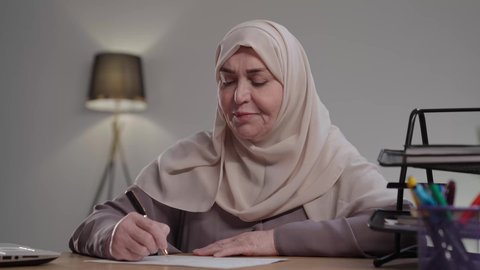 استخدام الاوراق البيضاء الفارغة في العمل، إدارة الأعمال وتنظيمها،تدوين وتسجيل الملاحظات والمعلومات على الورق,  تصوير لامراه عربيه خليجيه سعوديه كبيره بالسن ترتدي العباءه والحجاب تجلس على مكتب ، وتقوم بكتابة المهام والأهداف اليومية