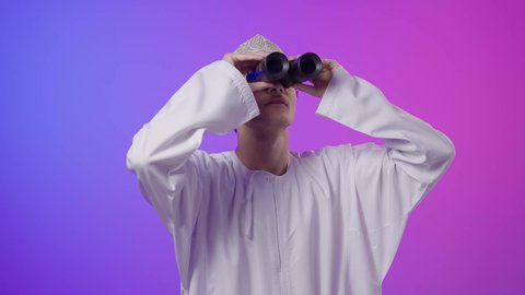 حب الاستكشاف و المغامرة، إيماءات الذهول والتفاجؤ, بورتريه مقرب لولد عربي خليجي عماني يرتدي الدشداشة و الكمة يحمل بيده منظار ثنائي العينين ينظر إلى شيئ ما, مفهوم النظرة المستقبلية، رؤية الاشياء البعيدة عبر التلسكوب، خلفية ملونه