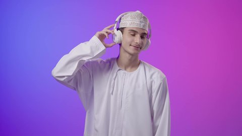 تسليه وترفيه، قضاء أوقات ممتعة بالاستماع للمواهب الغنائية، استخدام تقنية الهيدفون، بورترية لولد عربي خليجي عماني يرتدي الدشداشة و الكمة يضع سماعة الرأس يقوم بالإصغاء والاستماع إلى شيء ما، ايماءات وجه ويدين تدل على السعاده والتفاعل مع الأغاني, خلفية ملونه