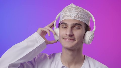 الاستمتاع والمرح، ايماءات وجه ويدين تدل على السعاده والتفاعل مع الأغاني، استخدام تقنية الهيدفون، قضاء أوقات ممتعة بالاستماع للمواهب الغنائية، بورترية مقرب لولد عربي خليجي عماني يرتدي الدشداشة و الكمة يضع سماعة الرأس يقوم بالإصغاء والاستماع إلى شيء ما، خلفية ملونه