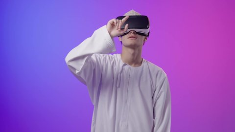 محاكاة العالم الخيالي، الاشارة باصبع السبابة إلى شيء ما، المتعة بالمشاهدة عبر نظارة الواقع الافتراضي، بورتريه لولد عربي خليجي عماني يرتدي الدشداشه والكمه ينظر من خلال نظارات ثلاثية الأبعاد (الفي ار)، استخدام جهاز تقني حديث ومتطور،خلفية ملونه