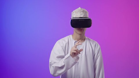 الاشارة باصبع السبابة إلى شيء ما، المتعة بالمشاهدة عبر نظارة الواقع الافتراضي، بورتريه لولد عربي خليجي عماني يرتدي الدشداشه والكمه ينظر من خلال نظارات ثلاثية الأبعاد (الفي ار)، محاكاة العالم الخيالي، استخدام جهاز تقني حديث ومتطور،خلفية ملونه