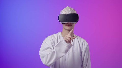 المتعة بالمشاهدة عبر نظارة الواقع الافتراضي، بورتريه لولد عربي خليجي عماني يرتدي الدشداشه والكمه ينظر من خلال نظارات ثلاثية الأبعاد (الفي ار), الاشارة باصبع السبابة إلى شيء ما، محاكاة العالم الخيالي، استخدام جهاز تقني حديث ومتطور،خلفية ملونه