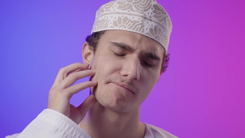 التهاب اللثه وتسوس الأَضراس, الاحتياج الى زيارة طبيب الأسنان، بورتريه مقرب لولد عربي  خليجي عماني يرتدي الدشداشة و الكمه يضع يده على خده بإيماءات تدل على ألم الأسنان,  التعب والإرهاق والحاجة لطلب العلاج والمساعده, خلفية ملونه