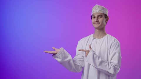 ايماءات التأكد من شيء ما ,التحديد نحو هدف معين, بورتريه لولد عربي اماراتي خليجي عماني يرتدي الدشداشة و الكمة يرفع اصبع السبابة للاعلى بتعابير السرور, الإشارة الى شخص ما, خلفية ملونه