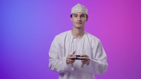 قضاء أوقات ممتعة في ألعاب الفيديو الترفيهيه، بورتريه لولد عربي خليجي عماني يرتدي الدشداشة والكمة يحمل بيده عصا التحكم، إيماءات الاستمتاع والاندماج، استخدام الأجهزة التكنولوجية والتقنيات الحديثة، خلفية ملونه