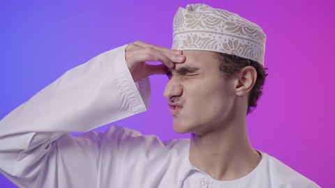 الشعور بالإرهاق وفقدان التركيز، الاجهاد والضغط النفسي, الاحساس بالشقيقية والصداع النصفي, بورتريه مقرب لولد عربي خليجي عماني يرتدي الدشداشة و الكمه يضع يديه على رأسه ويغمض عينيه بإيماءات ألم الرأس الشديد, خلفية ملونه