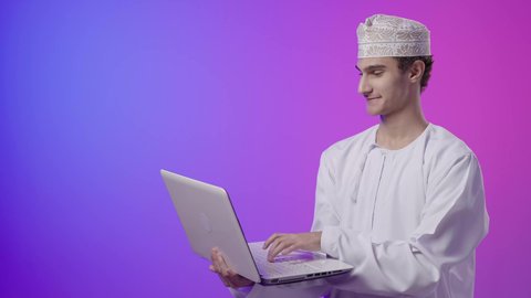 Entertainment and leisure by spending time browsing social media on a laptop, following lessons and tutorials remotely, a portrait of an Arab Gulf Omani boy wearing a dishdasha and kumma, smiling while using a laptop, using technological devices in learning, colorful background.