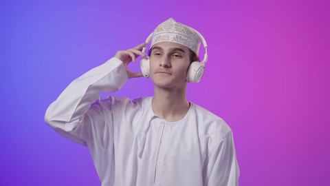Facial and hand gestures indicating happiness and interaction with songs, use of headphone technology, spending enjoyable times listening to singing talents, a portrait of an Arab Gulf Omani boy wearing a dishdasha and kumma, wearing headphones and listening to something, enjoyment and fun, colorful background.
