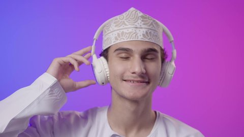 Using headphone technology, spending enjoyable times listening to singing talents, a close-up portrait of an Omani Gulf Arab boy wearing a dishdasha and kumma, putting on headphones, listening to something, facial and hand gestures indicating happiness and interaction with the songs, enjoyment and fun, colorful background.