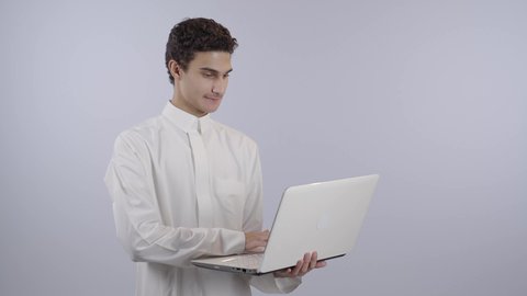 الكتابة على لوحة المفاتيح، حل الواجبات المنزليه عبر اللابتوب، ادخال التقنية والتكنولوجيا بالتعليم، بورتريه لولد عربي خليجي سعودي يرتدي الثوب الابيض يقوم بمتابعة منصات التعليم الالكتروني المتاحة عبر الانترنت بإستخدام جهاز الحاسب المحمول، النظر الى الكاميرا بايماءات السعادة و السرور، خلفية بيضاء