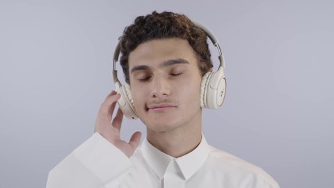 سماع الموسيقى والصوتيات، التسلية والترفيه بإستخدام تقنية الهيدفون، ارتداء سماعات الرأس الصوتية، بورتريه مقرب لولد عربي خليجي سعودي يرتدي الثوب الابيض يضع يقوم بالاستماع للأغاني بايماءات السرور والسعادة ، خلفية بيضاء