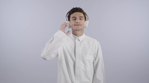 التسلية والترفيه بإستخدام تقنية الهيدفون، سماع الموسيقى والصوتيات، ارتداء سماعات الرأس الصوتية، بورتريه لولد عربي خليجي سعودي يرتدي الثوب الابيض يضع يقوم بالاستماع للأغاني بايماءات السرور والسعادة ، خلفية بيضاء
