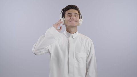 التفاعل مع الأنغام الموسيقية باستخدام الهيدفون، إيماءات الاندماج مع الأغنية، بورتريه لولد عربي خليجي سعودي يرتدي الثوب الابيض يستمع إلى الموسيقى من خلال سماعة الرأس بتعابير الانصات والاستمتاع، موهبة غنائية فنية،خلفية بيضاء