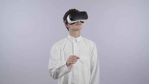 المتعة والتسلية بالمشاهدة عبر نظارة الواقع الافتراضي، محاكاة العالم الخيالي، استخدام جهاز تقني حديث ومتطور، بورتريه لولد عربي خليجي سعودي يرتدي الثوب الابيض ينظر من خلال نظارات ثلاثية الأبعاد (الفي ار) بإيماءات الاندماج,، خلفيه بيضاء