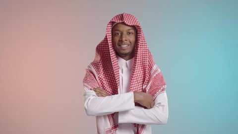 A portrait of a smiling young Arab Gulf Saudi man with a dark complexion, wearing traditional thobe and shemagh, standing with arms crossed, exuding self-confidence and a masculine appearance, with attention to external appearance, against a colorful background.