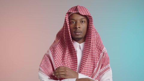 Standing upright and confidently, paying attention to appearance, a young dark-skinned Arab Gulf man wearing traditional thobe and ghutrah stands with crossed arms, against a colorful background.