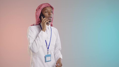 اجراء مكالمه هاتفيه اثناء العمل, دمج الاجهزه والتقنيات الحديثه في العمل, موظف عربي خليجي سعودي يرتدي الثوب والشماغ التقليدي وبطاقه تعريفيه زرقاء يقوم بماكلمه هاتفيه, خلفيه ملونه