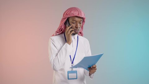 An Arab Gulf Saudi employee wearing traditional thobe and shemagh is using a mobile phone to follow up on work while holding documents in his hand, using modern devices and technologies, with a colored background.