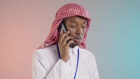 موظف عربي خليجي سعودي يرتدي الثوب والشماغ التقليدي يستخدم الهاتف المحمول لمتابعة العمل يحمل مستندات في يده, استخدام الاجهزه والتقنيات الحديثه, خلفيه ملونه