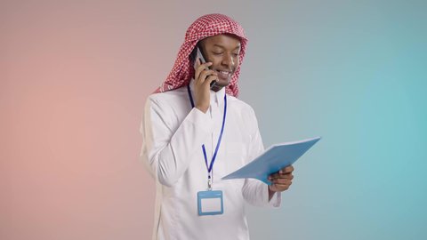 A Saudi Arabian Gulf man wearing traditional thobe and shemagh is using a mobile phone to follow up on work while holding documents in his hand, using modern devices and technologies, with a colorful background.