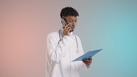 المناقشه في العمل حول التقارير الطبيه, طبيب عربي خليجي سعودي يرتدي المعطف والسماعه الطبيه يقوم باجراء مكالمه هاتفيه لمراجعه النتائج مع شخص ما, خلفيه ملونه