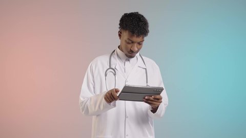 استخدام احدث الاجهزه والتقنيات الحديثه, دمج الاجهزه الحديثه في المجال الطبي, طبيب عربي خليجي سعودي يرتدي المعطف الابيض والسماعه الطبيه يقوم باستخدام جهاز التابلت لفحص التقارير الطبيه, خلفيه ملونه
