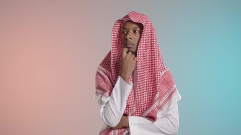 Thinking and pondering over something, a young Arab Gulf Saudi man wearing traditional thobe and shemagh is placing his hand on his face to contemplate something, with a colorful background.