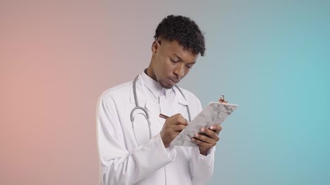 وصف العلاج المناسب للمرضي, طبيب عربي خليجي سعودي يرتدي المعطف الابيض والسماعه الطبيه يقوم بكتابه التقارير الطبيه في سجل الملاحظات,  خلفيه ملونه