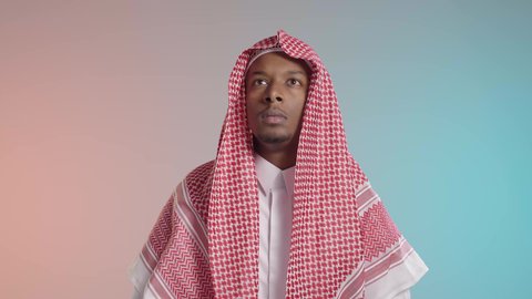 Contemplation about daily matters, a young Arab Gulf Saudi man wearing traditional thobe and shemagh looking up with a gesture of confusion and contemplation, colorful background.