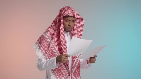 المقارنه والتمعن في قرآءه النتائج, شاب عربي خليجي سعودي يرتدي الثوب والشماغ التقليدي يقوم بمراجعه الاوراق, ايماءات القلق والتوتر, خلفيه ملونه
