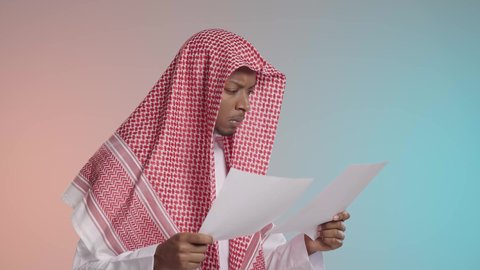 التمعن في قرآءه النتائج والمقارنه بينهم, شاب عربي خليجي سعودي يرتدي الثوب والشماغ التقليدي يقوم بمراجعه الاوراق, ايماءات القلق والتوتر, خلفيه ملونه
