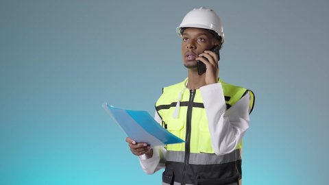 استخدام جهاز تقني حديث متطور، مفهوم الهندسة والإنشاء المعماري, إجراء مكالمة صوتية، مهندس عربي خليجي عماني يرتدي سترة عاكسة و خوذة بيضاء و يحمل ملف ملاحظات