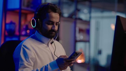 Browsing the internet via mobile phone, using advanced editing software to produce videos, meditation and artistic creativity in front of the screen, filming an Arab Gulf Saudi editor wearing a white thobe and headphones conducting visual analysis.