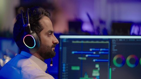 مفهوم الإبداع في صناعة المحتوى المرئي، التأمل والإبداع الفني أمام الشاشة، تصوير مقرب من الجانب لمونتير عربي خليجي سعودي يرتدي الثوب الابيض و سماعات الرأس يقوم التفكير العميق والتحليل البصري