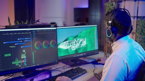 ايماءات السعادة و السرور، الاستفادة من برامج وتقنيات المونتاج الحديثة، تصوير من الجانب لمونتير عربي خليجي سعودي يرتدي الثوب الابيض  يضع سماعات الرأس ويقوم بتعديل الصوت على شاشة الكمبيوتر، صناعة الأفلام والبرامج التلفزيونية