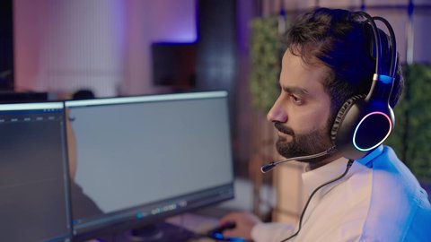 مفهوم الإبداع والإخراج الفني، تصويرمقرب من الجانب لمونتير عربي خليجي سعودي يرتدي الثوب الابيض و سماعات الرأس يجلس أمام جهاز الحاسوب يستخدم برامج المونتاج لإضافة المؤثرات البصرية والصوتية