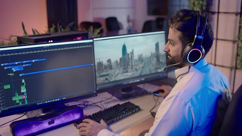 النقر على لوحه المفاتيح، استخدام التقنية الحديثة، الابتكار والإبداع في تصميم وإنتاج المحتوى المرئي، تصويرمن الجانب لرجل عربي خليجي سعودي يرتدي الثوب الابيض يضع سماعة الراس يعمل في غرفة المونتاج يستخدم الحاسب المحمول لقص وترتيب المشاهد، ايماءات الاندماج و التركيز
