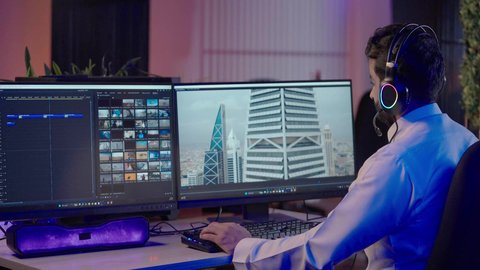 ايماءات السعادة و السرور، استخدام التقنية الحديثة، الابتكار والإبداع في تصميم وإنتاج المحتوى المرئي، تصويرمن الجانب لرجل عربي خليجي سعودي  يرتدي الثوب الابيض يضع سماعة الراس يعمل في غرفة المونتاج يستخدم الحاسب المحمول لقص وترتيب المشاهد