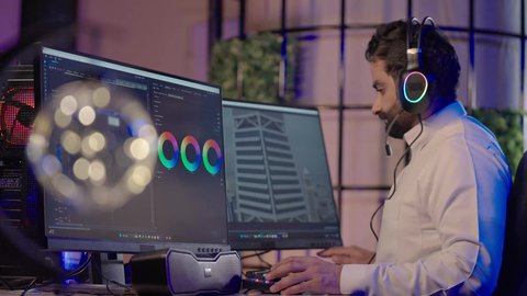 استخدام التقنية الحديثة، ايماءات الاندماج و التركيز، الابتكار والإبداع في تصميم وإنتاج المحتوى المرئي، تصويرمن الجانب لرجل عربي خليجي سعودي  يرتدي الثوب الابيض يضع سماعة الراس يعمل في غرفة المونتاج يستخدم الحاسب المحمول لقص وترتيب المشاهد يضع يده على ذقنه للتفكير فى شيء ما