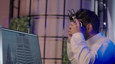 تقنيات الاتصال الحديثة في بيئة العمل الرقمية، تصويرمقرب من الجانب لرجل عربي خليجي سعودي  يرتدي الثوب الابيض يضع سماعة الراس يعمل في غرفة المونتاج يستخدم الحاسب المحمول لقص وترتيب المشاهد، مفهوم صناعة المحتوى المرئي والإبداع الفني