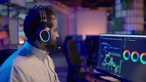 مفهوم الإبداع والإخراج الفني، تصوير من الجانب لرجل عربي خليجي سعودي يرتدي الثوب الابيض و سماعات الراس يعمل في غرفة المونتاج يستخدم شاشة متعددة لقص وترتيب المشاهد، مفهوم صناعة المحتوى المرئي والإبداع الفني، ايماءات الاندماج و التركيز