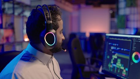 بيئة عمل تقنية حديثة متعددة الشاشات، تصوير من الجانب لمونتيرعربي خليجي سعودي يرتدي الثوب الابيض و سماعات الراس يجلس أمام شاشة حاسب محمول يقوم باخراج مشهد سينمائي  ، ايماءات الاندماج و التركيز