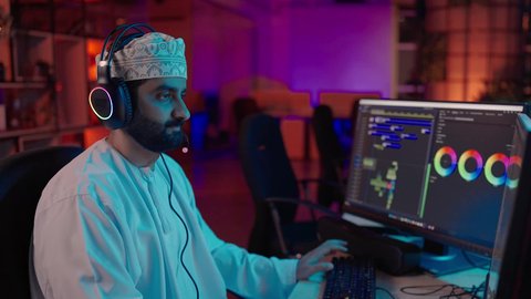 التحكم في الانتقالات والمؤثرات، تصويرمن الجانب لمونتير عربي خليجي عماني يرتدي الدشداشة و الكمة وسماعات الرأس يجلس داخل الاستديو يعمل على ترتيب المشاهد باستخدام برامج الحاسوب المتطورة ، ايماءات التفكير و التركيز