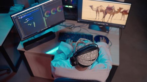 مفهوم صناعة المحتوى الرقمي، تصوير من الخلف لمونتير عربي خليجي عماني يرتدي الدشداشة و الكمة وسماعات الرأس يجلس داخل الاستديو يعمل على ترتيب المشاهد باستخدام برامج الحاسوب المتطورة ، ايماءات التفكير و التركيز