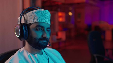 مونتاج وإخراج مرئي، تصوير مقرب لمونتير عربي خليجي عماني يرتدي الدشداشة و الكمة وسماعات الرأس يجلس داخل الاستديو يعمل على ترتيب المشاهد باستخدام برامج الحاسوب المتطورة ، ايماءات التفكير و التركيز