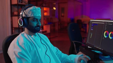 الإبداع في مجال صناعة المحتوى، تصوير لمونتير عربي خليجي عماني يرتدي الدشداشة و الكمة وسماعات الرأس يجلس داخل الاستديو يعمل على ترتيب المشاهد باستخدام برامج الحاسوب المتطورة ، ايماءات التفكير و التركيز